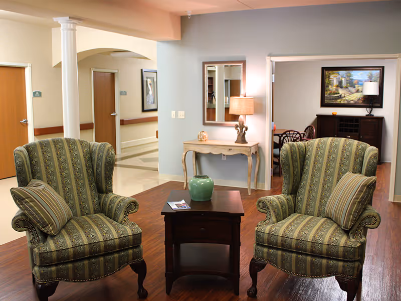 A cozy seating area in a senior living facility with two patterned armchairs facing each other and a small wooden table with a green vase between them. In the background, there is a hallway with doors, a console table with a lamp and decorative items, a mirror on the wall, and a framed painting above a cabinet.