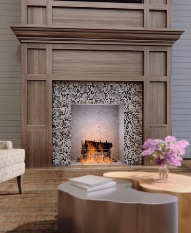 Cozy living room with a lit fireplace set in a decorative wooden mantel, a coffee table with books and a vase of pink flowers, and part of an upholstered chair.