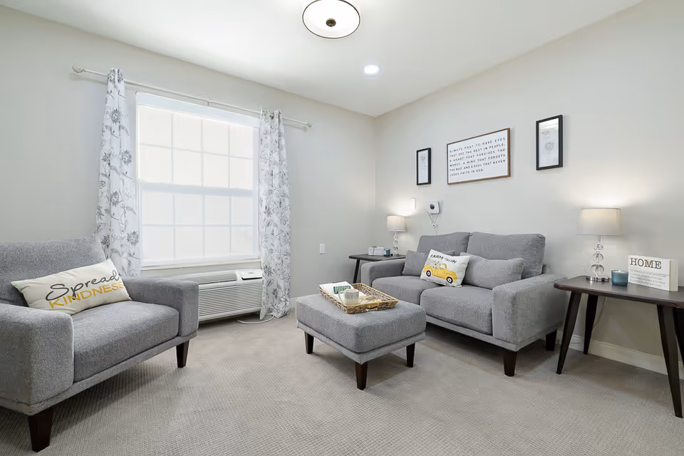 A cozy living room with light gray walls and carpet. The room features a gray armchair with a 'Spread Kindness' pillow, a matching gray loveseat with decorative pillows, and a gray ottoman with a tray on top. There are two side tables with lamps and decorative items, a large window with floral curtains, and framed artwork on the wall.