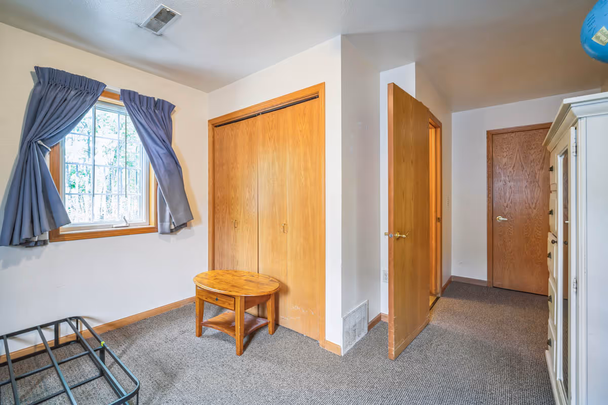 A small bedroom with a window covered by blue curtains, a wooden closet with double doors, a small round wooden table, an open wooden door leading to a hallway, and a white dresser on the right side.