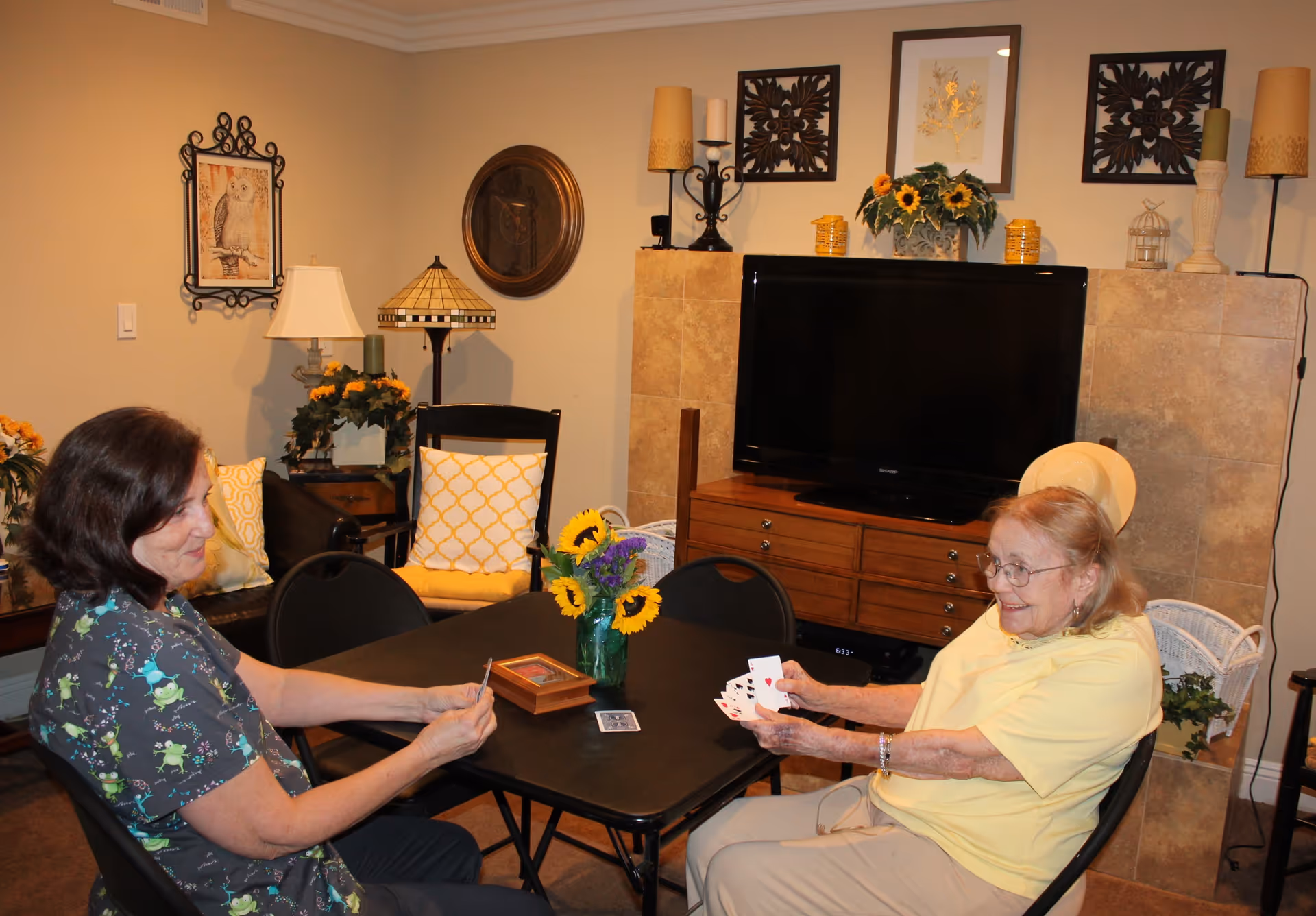 Two women sitting at a black table playing cards in a cozy living room. The room features a large flat-screen TV on a wooden cabinet, decorative wall art, lamps, and yellow-themed cushions and flowers.