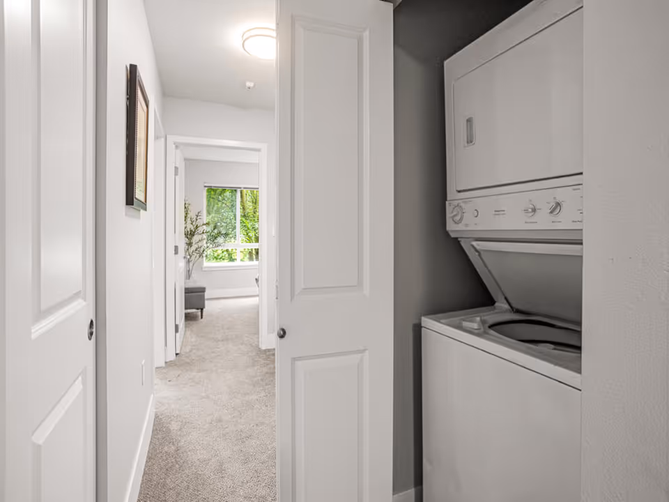 View down a carpeted hallway with white walls and doors, featuring a stacked washer and dryer unit on the right side behind a partially open door. At the end of the hallway, there is a room with a large window showing green trees outside and a small gray bench with a plant beside it.