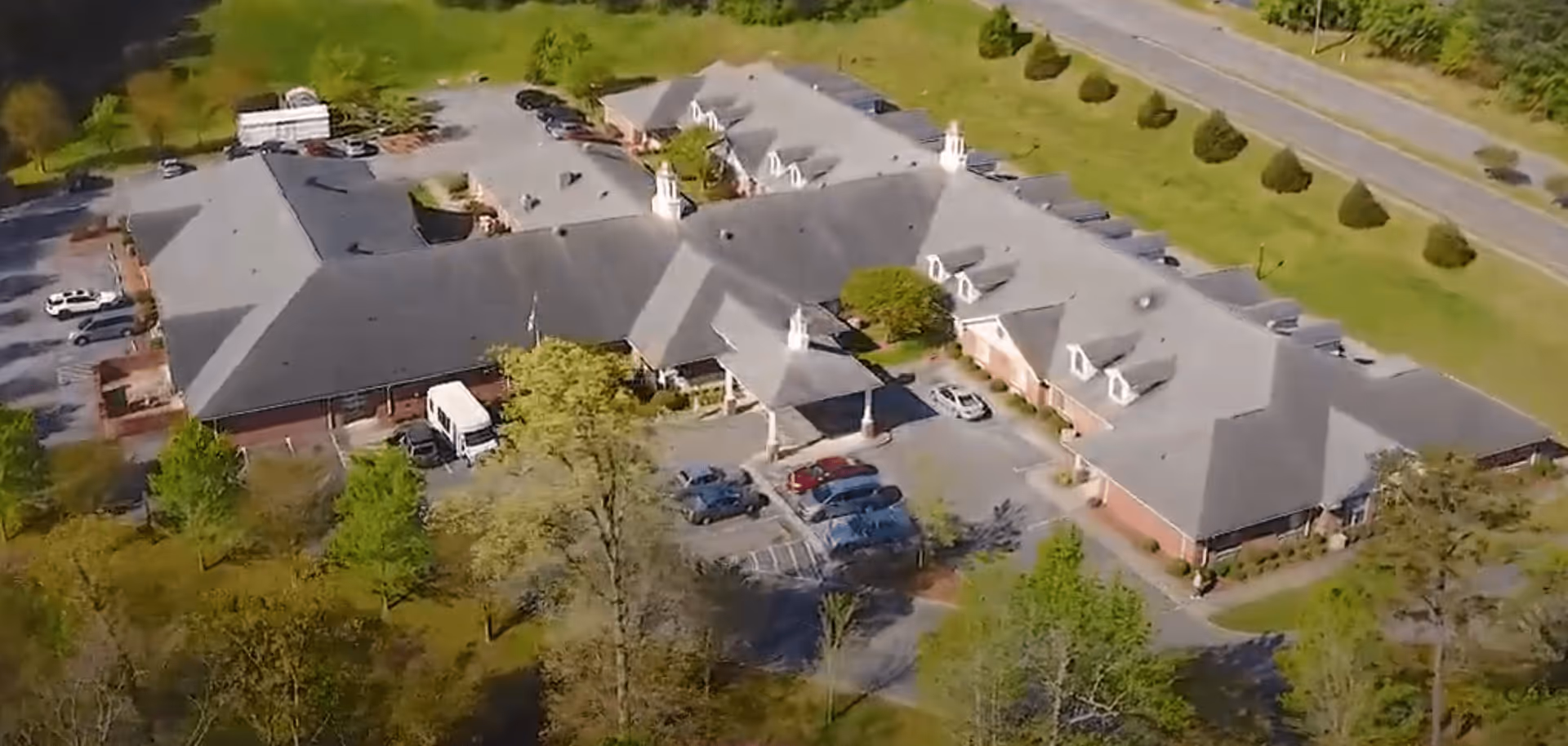 Aerial view of a one-story assisted living building with multiple roof sections, a central entrance and parked cars, surrounded by trees and lawns.