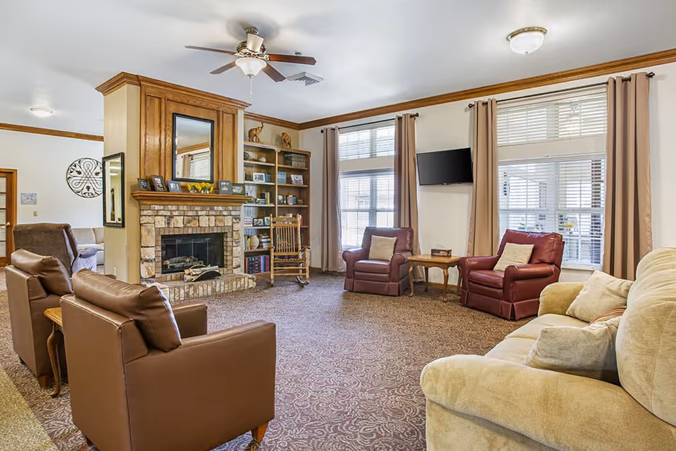A cozy living room with a stone fireplace and wooden mantel, flanked by built-in shelves filled with books and decorative items. The room features two brown leather armchairs, two red leather armchairs with pillows, a beige sofa with pillows, a wooden rocking chair, and a small wooden side table. Large windows with beige curtains allow natural light to fill the space, and a ceiling fan with a light fixture is mounted on the ceiling. A flat-screen TV is mounted on the wall between the windows.