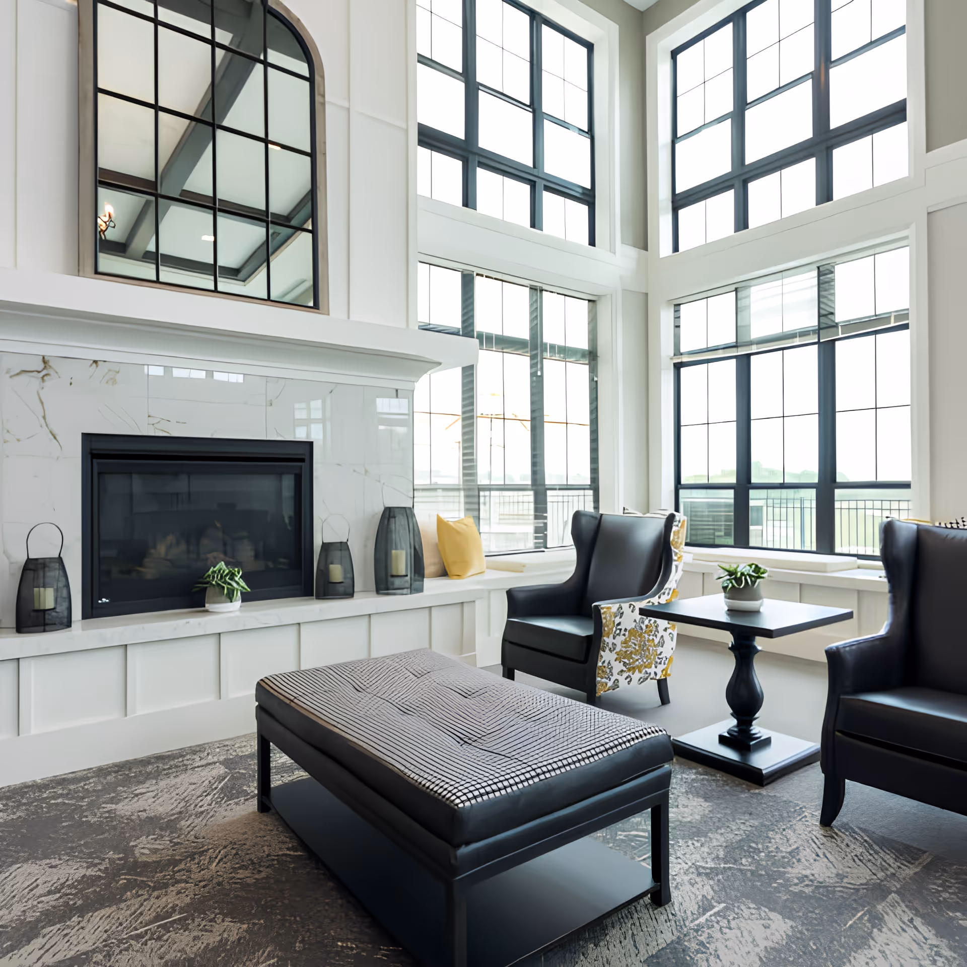 A bright and spacious senior living facility common area with large floor-to-ceiling windows, two black leather armchairs with floral patterned sides, a black cushioned ottoman, a small black side table with a potted plant, and a modern fireplace with decorative lanterns and a large window above it.