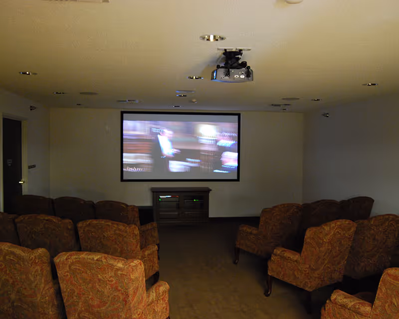 Small screening room with rows of patterned armchairs facing a wall-mounted projection screen and a ceiling projector.