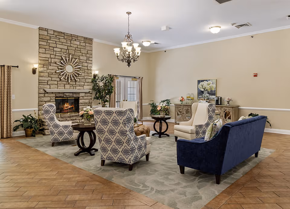 A cozy living room area with a stone fireplace, a sunburst mirror above it, and a lit fire inside. The room features a blue sofa, two patterned armchairs, a beige armchair, and two round wooden tables with flower arrangements. There is a chandelier hanging from the ceiling, plants in the corners, and a sideboard with decorative items and a painting on the wall. The floor is a combination of wood and a large area rug.