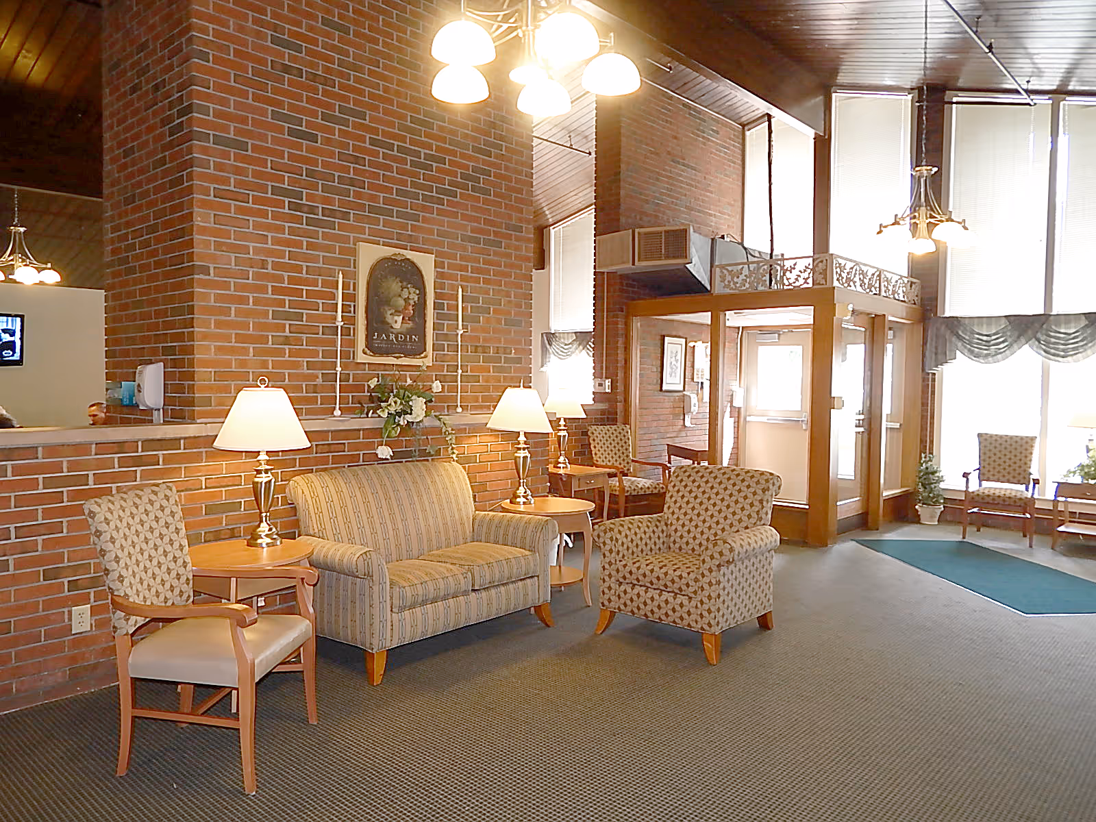A cozy seating area in a senior living facility lobby with patterned armchairs, a striped loveseat, wooden side tables with lamps, and a brick wall with a framed floral picture. Large windows with blinds and valances let in natural light, and a glass entryway door is visible in the background.