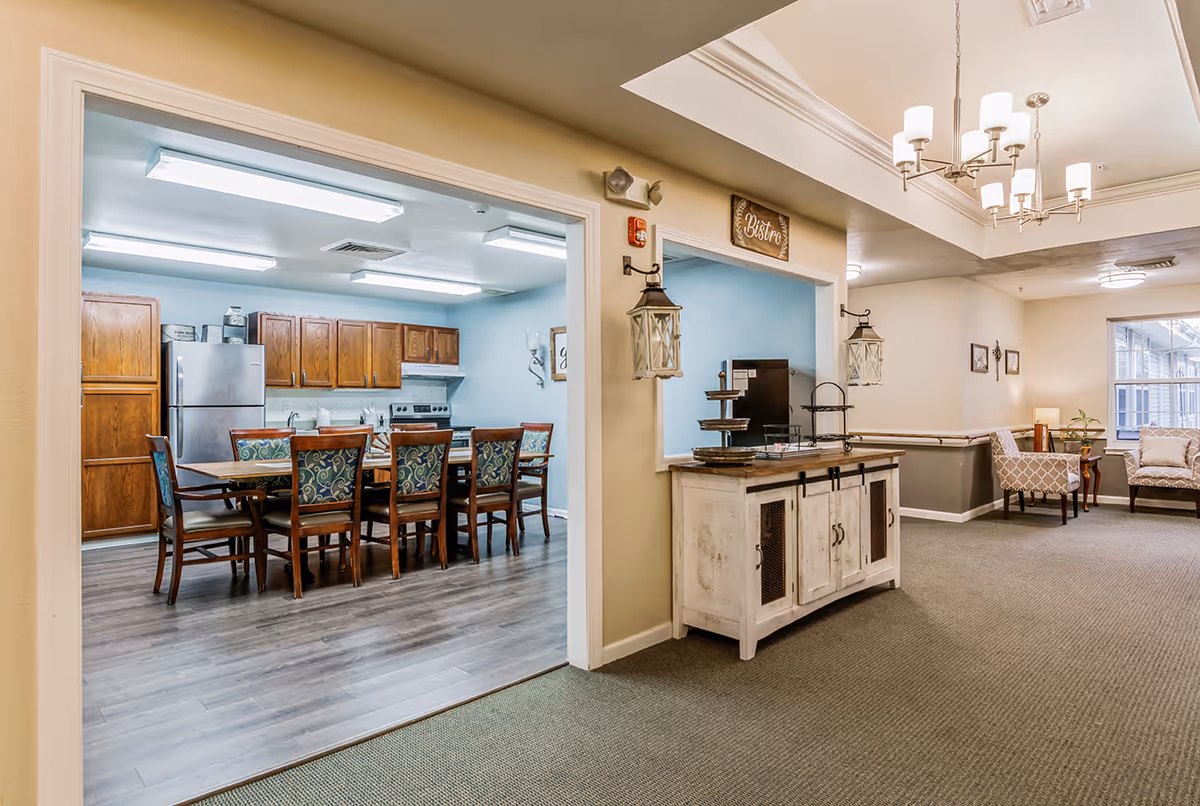 Interior view of a senior living facility showing a dining area with a wooden table and chairs with patterned cushions, adjacent to a kitchen with wooden cabinets, a refrigerator, and an oven. The foreground features a hallway with carpeted flooring, a white sideboard with decorative lanterns, and a seating area with armchairs and a window letting in natural light.