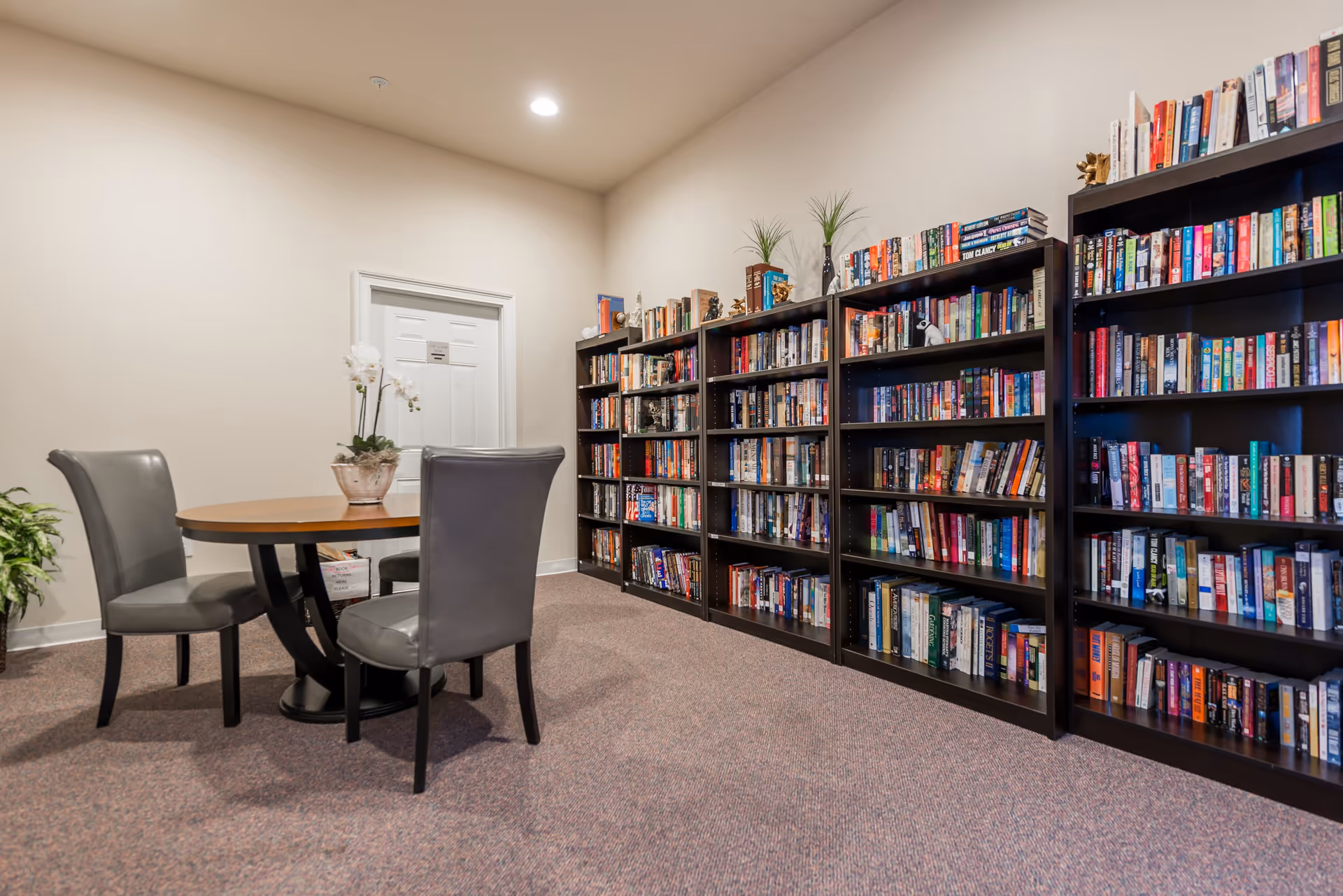 Interior room with a round wooden table surrounded by three gray chairs. Along one wall are several tall black bookshelves filled with books and decorated with small plants and figurines. The room has beige walls and carpeted floor, with a closed white door in the background.