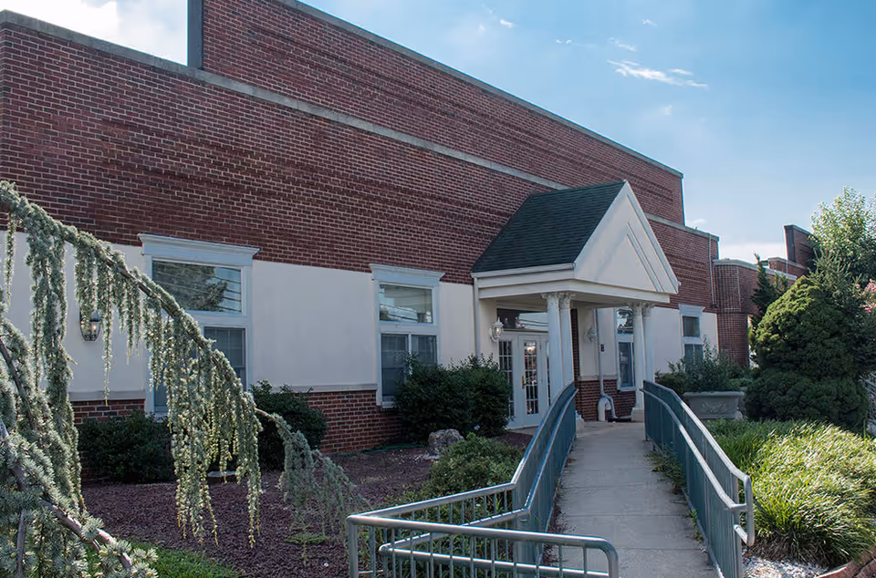 Entrance of a brick building with a covered portico, wheelchair ramp, and surrounding shrubs under a blue sky.