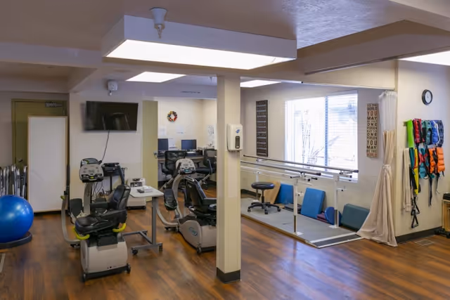 Interior view of a rehabilitation and care center room with exercise equipment including stationary bikes, parallel bars for walking practice, therapy balls, and various physical therapy tools mounted on the wall. The room has wood flooring, a large window with blinds, and a wall-mounted TV.