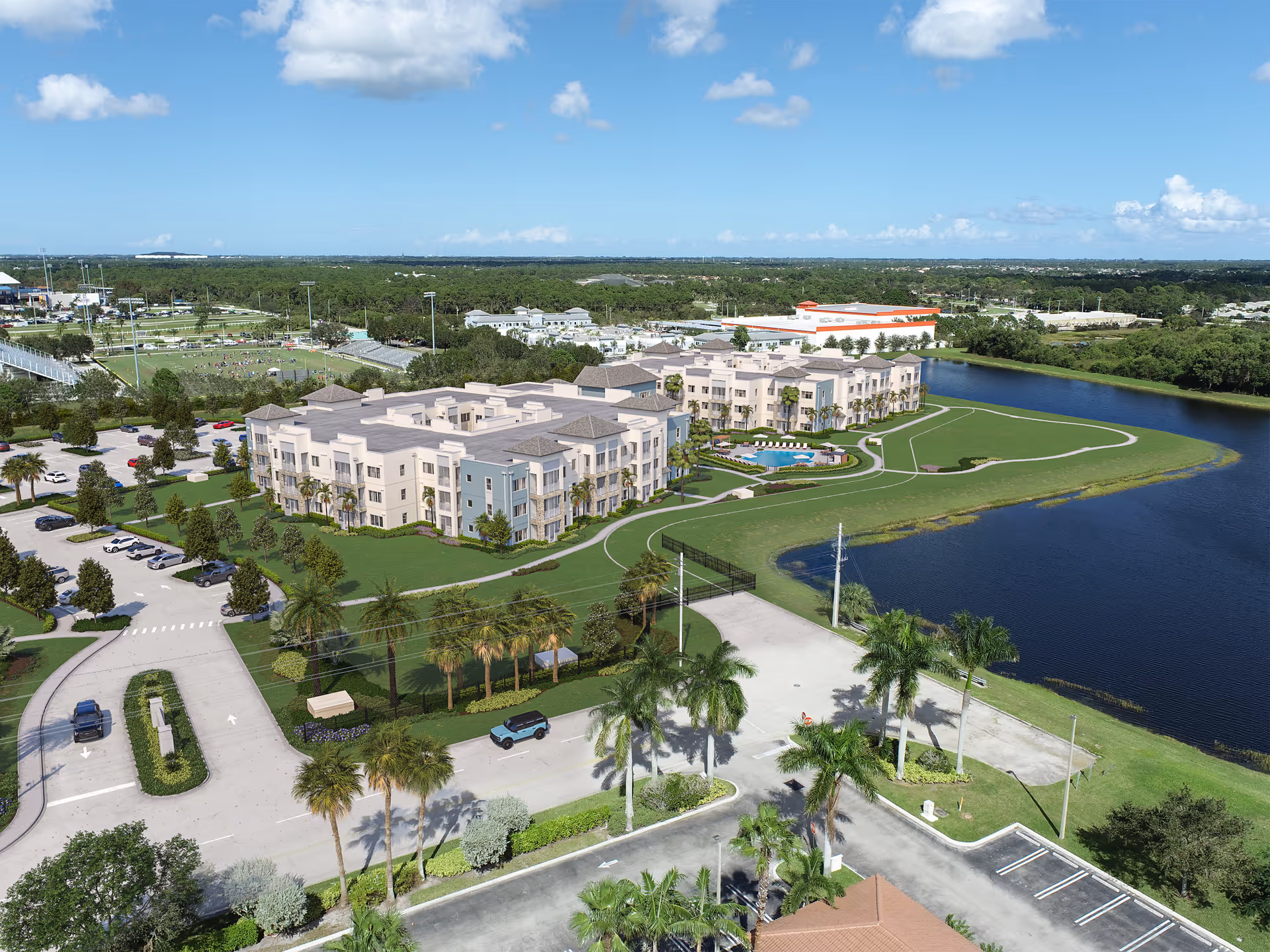 Aerial view of AVIVA Port St. Lucie West senior living facility showing multiple beige and light blue residential buildings surrounded by green lawns, palm trees, a parking lot with cars, walking paths, and a large body of water on the right side under a blue sky with scattered clouds.