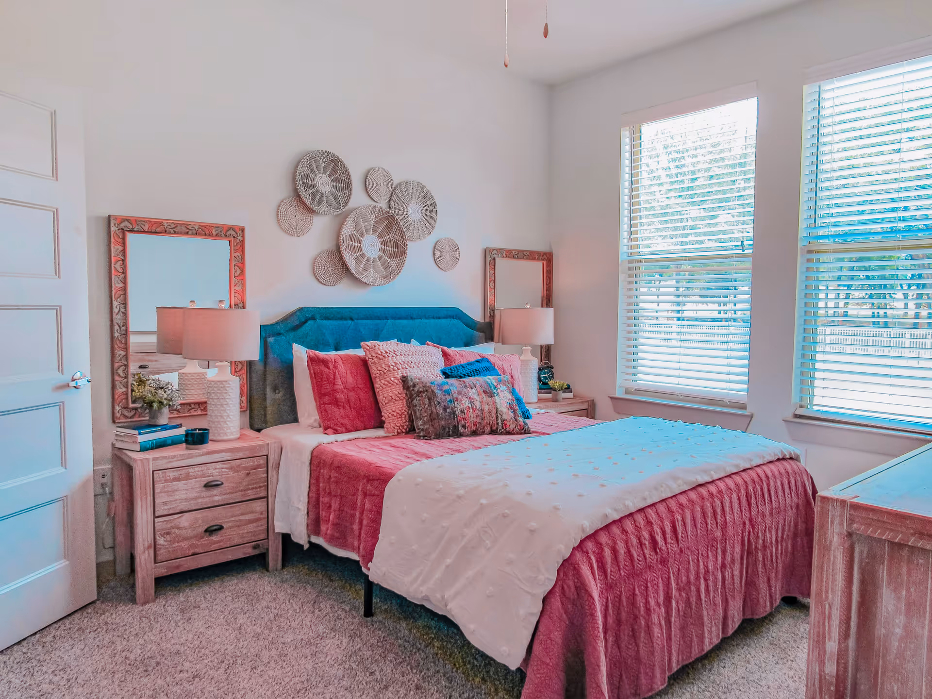 A bright bedroom with a bed featuring a teal upholstered headboard and pink bedding, including multiple pillows. On each side of the bed are wooden nightstands with white lamps and decorative items. Two large windows with white blinds allow natural light to fill the room. The walls are white with decorative woven wall hangings above the bed.