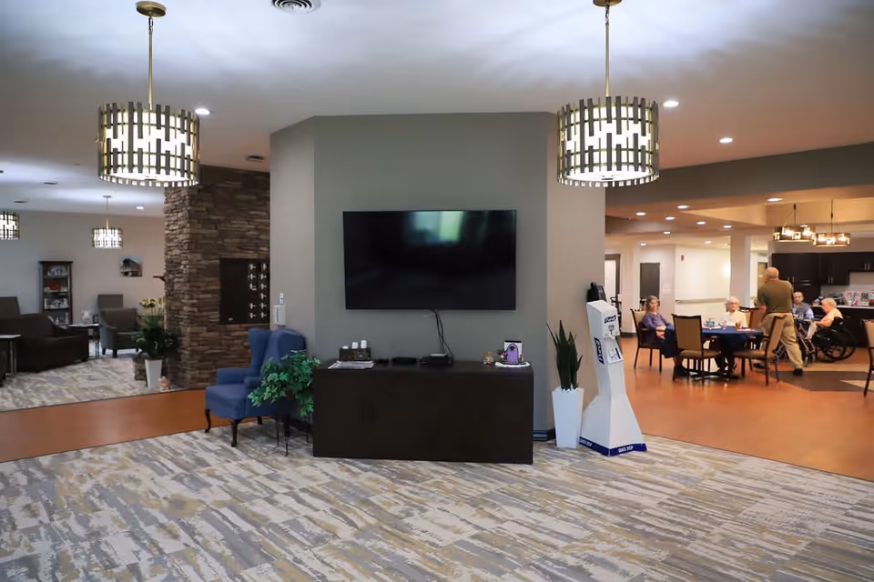 Interior view of a senior living facility common area with a mounted flat-screen TV on a gray wall, two blue armchairs, a dark wooden cabinet, and decorative plants. In the background, several elderly people are seated around a table in a dining area, with a staff member standing nearby. The space is well-lit with modern hanging light fixtures and has a mix of carpet and wood flooring.