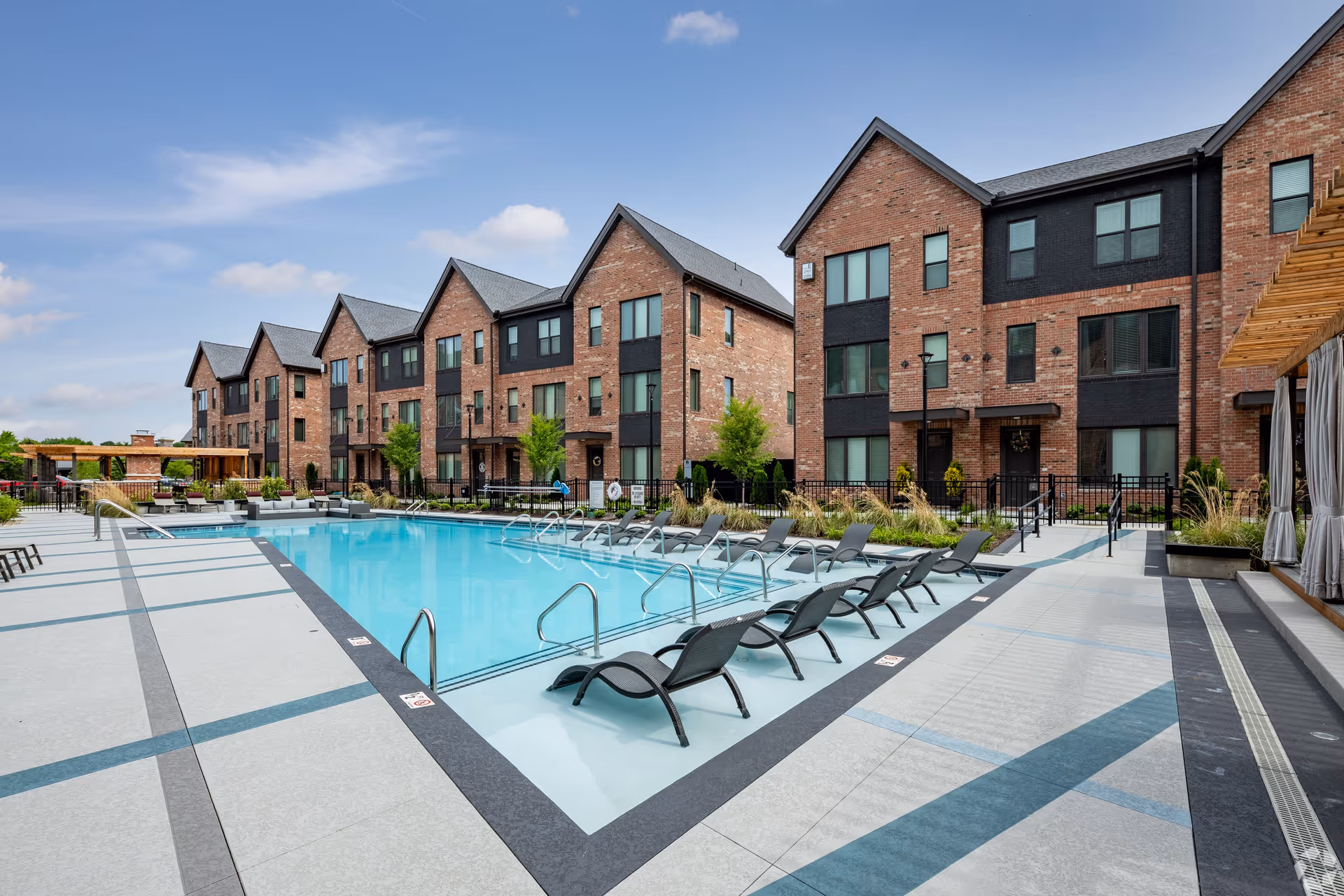 Outdoor swimming pool with lounge chairs partially submerged in the water, surrounded by a spacious pool deck. In the background, there are multiple three-story brick residential buildings with large windows and black accents. The sky is clear with a few clouds.