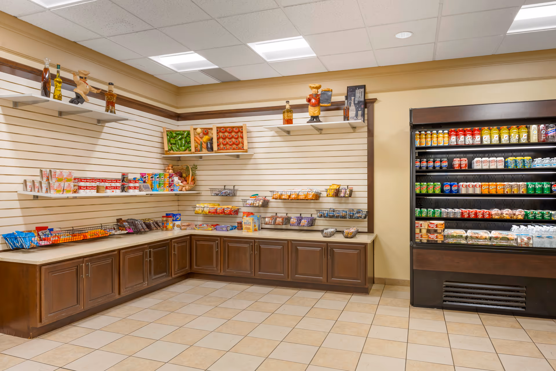 A small convenience store area inside a senior living facility with shelves stocked with snacks, canned goods, and beverages. There is a refrigerated display case on the right filled with various drinks and packaged food items. The walls have decorative shelves with bottles and figurines, and the floor is tiled.