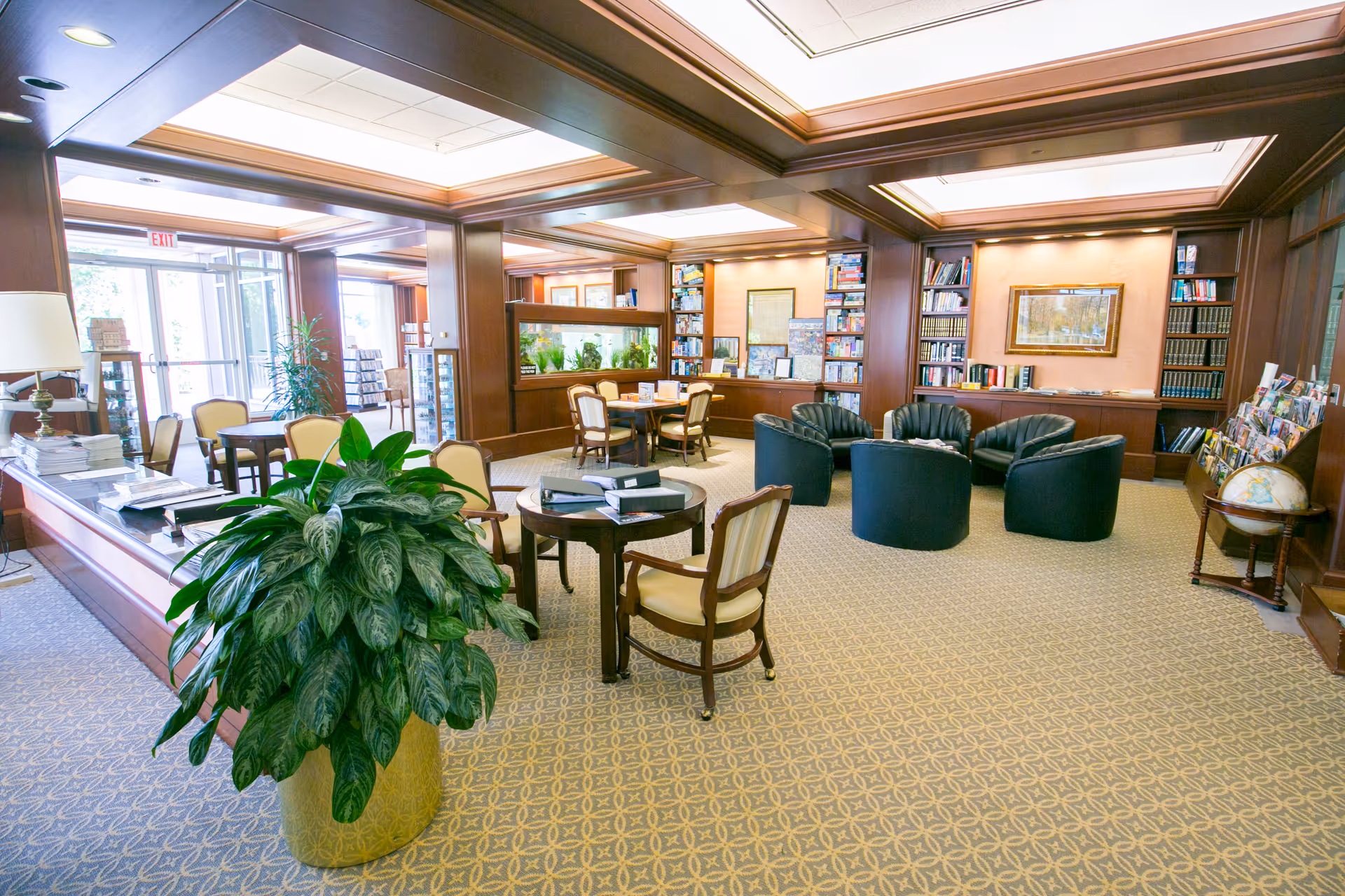 Bright wood-paneled common lounge with chairs, round tables, bookshelves, and large potted plants.
