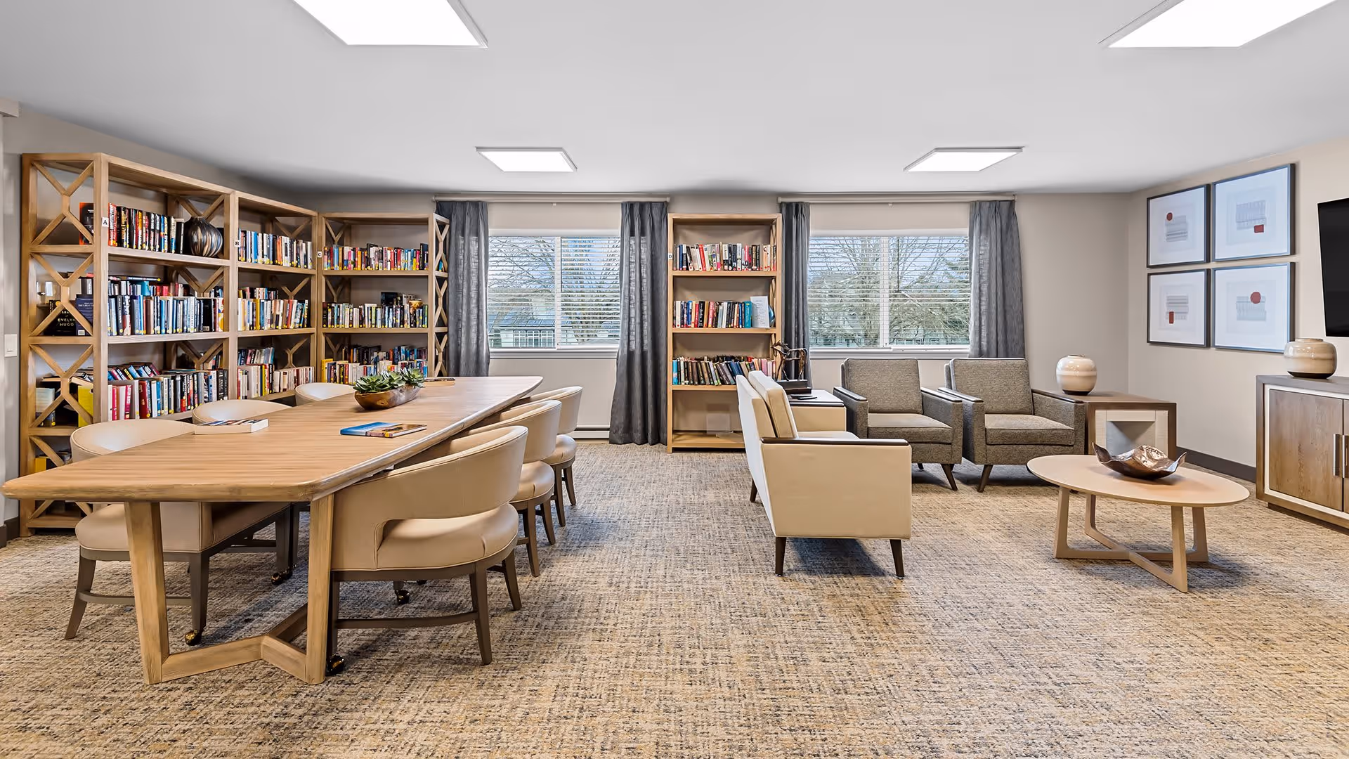 A bright and spacious common area with a long wooden table surrounded by beige chairs on the left, bookshelves filled with books along the back wall, and a seating area with beige and gray armchairs around a round wooden coffee table on the right. Large windows with gray curtains let in natural light, and framed artwork hangs on the wall above a wooden cabinet with a TV mounted beside it.