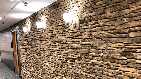 Interior hallway with a stacked-stone accent wall, three wall sconces, carpeted floor and a door at the end.