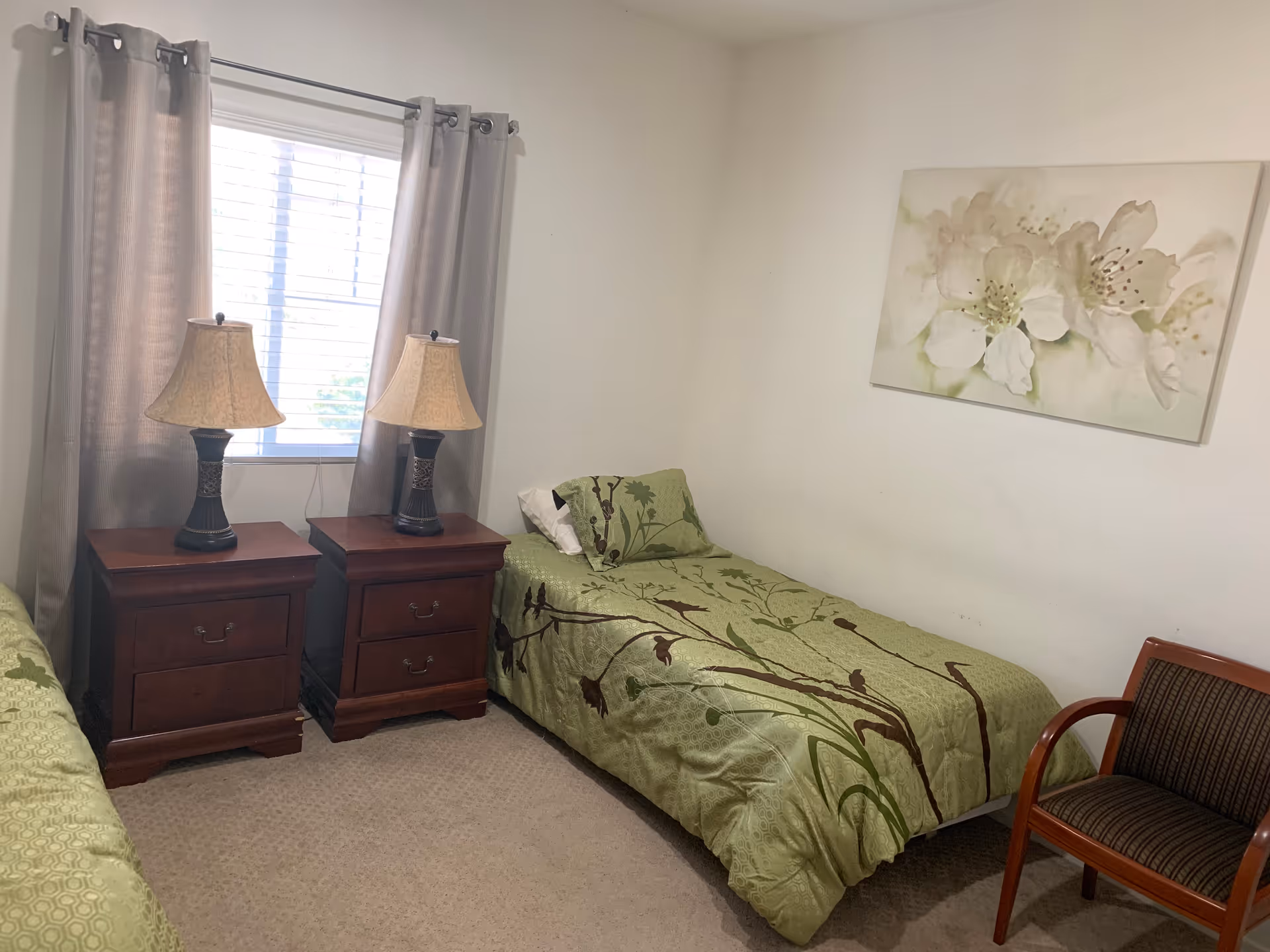 A small bedroom with two single beds covered in green bedspreads with a floral and bird pattern. Between the beds are two wooden nightstands, each with a lamp. A window with gray curtains is behind the nightstands. On the right side of the room is a wooden chair with striped upholstery. A large floral painting hangs on the white wall above one of the beds.