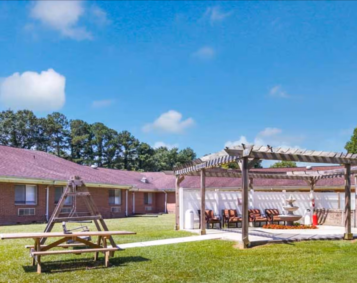 Brick building with purple roof surrounds grassy courtyard with a wooden pergola, fountain, and seating.