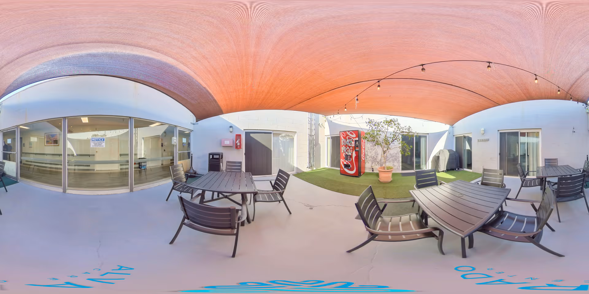 Covered outdoor courtyard with several tables and chairs, a vending machine, potted plant, and sliding glass doors into the building.