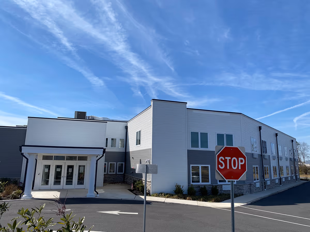 Exterior view of a modern two-story senior living facility building under a blue sky with some clouds. The building has white and gray siding with multiple windows and a covered entrance supported by white columns. In the foreground, there is a stop sign and some greenery near the parking area.