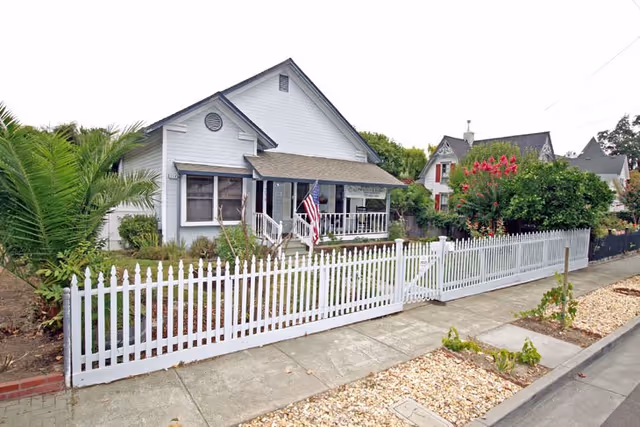 White cottage-style house with a front porch, American flag, and a white picket fence along the sidewalk.