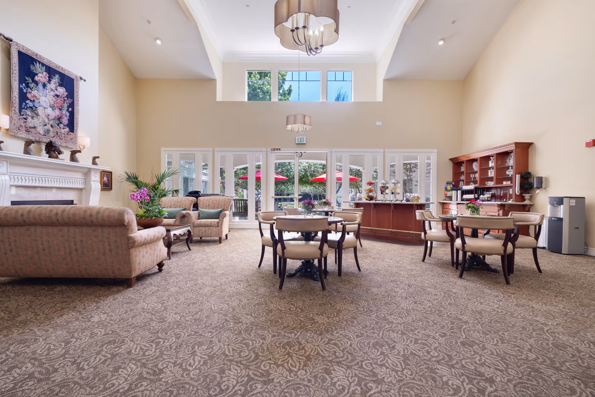 A spacious and well-lit common area with beige walls and carpet featuring a floral pattern. The room contains several upholstered armchairs and sofas arranged around a fireplace with a floral tapestry hanging above it. There are round tables with chairs in the center, decorated with small flower arrangements. A wooden cabinet with glassware and a counter with snacks and beverages is visible on the right side. Large windows and glass doors at the back open to an outdoor area with red umbrellas and greenery.