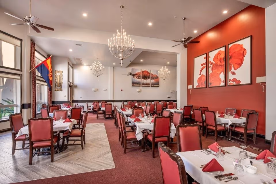 A spacious dining room with multiple tables covered in white tablecloths and set with red napkins, silverware, and glassware. The chairs have red upholstery and dark wooden frames. The room features chandeliers, ceiling fans, large windows with curtains, and red accent walls adorned with floral artwork.