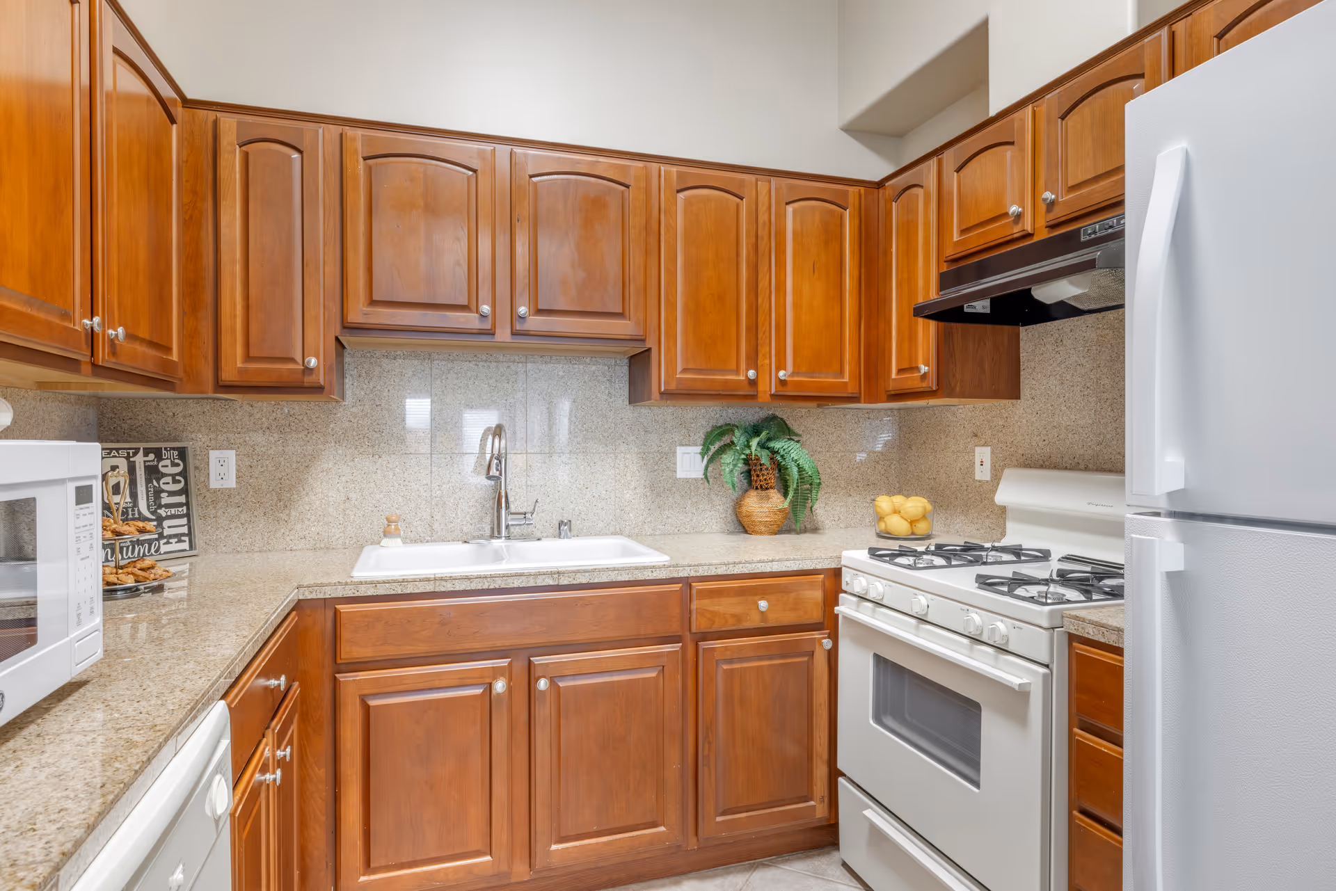 A kitchen with wooden cabinets, a white double sink, a white gas stove, a white refrigerator, a white microwave, and a beige granite countertop and backsplash. There is a small plant in a woven basket and a bowl of lemons on the counter.