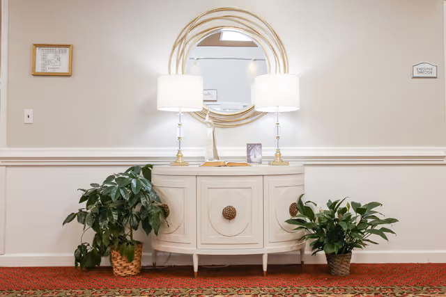 A hallway area with a white half-moon console table against a beige wall. On the table are two matching lamps with white shades, a small sculpture, an open book, and a framed photo. Above the table is a large round mirror with a gold frame. Two green potted plants are placed on the floor on either side of the table. The floor is covered with a red patterned carpet. A small sign on the wall to the right reads 'EXECUTIVE DIRECTOR'.