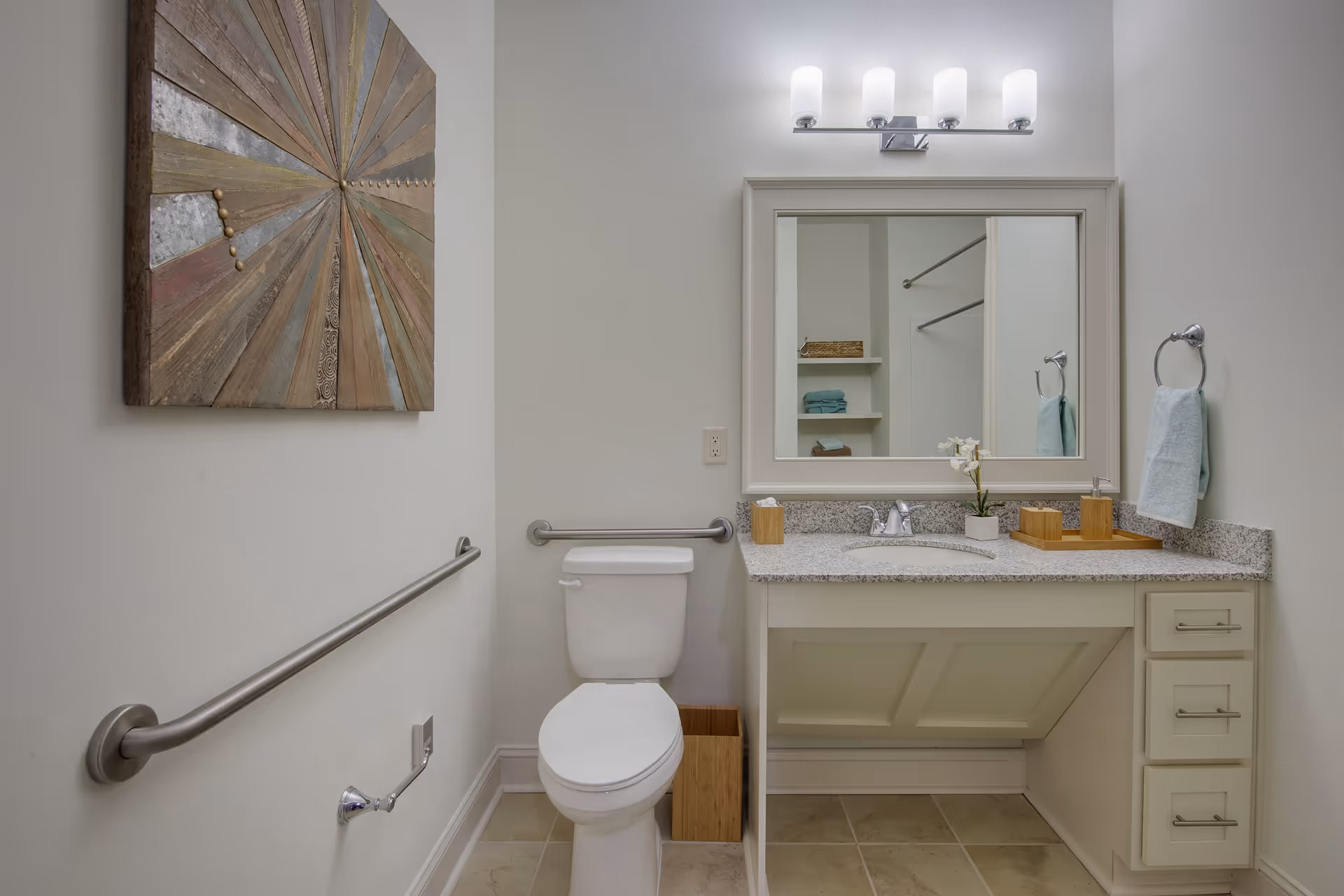 A clean and modern bathroom with a white toilet, a granite countertop with a sink, a large mirror above the sink, and a wall-mounted light fixture. There is a metal grab bar next to the toilet and a towel ring with a light blue towel. A piece of abstract wooden wall art is hung on the left wall.