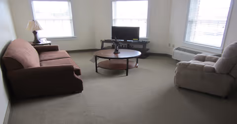 A living room with a brown sofa on the left, a beige recliner chair on the right, a round wooden coffee table in the center, and a small TV on a stand against the wall. The room has two windows with blinds and a lamp on a side table next to the sofa.