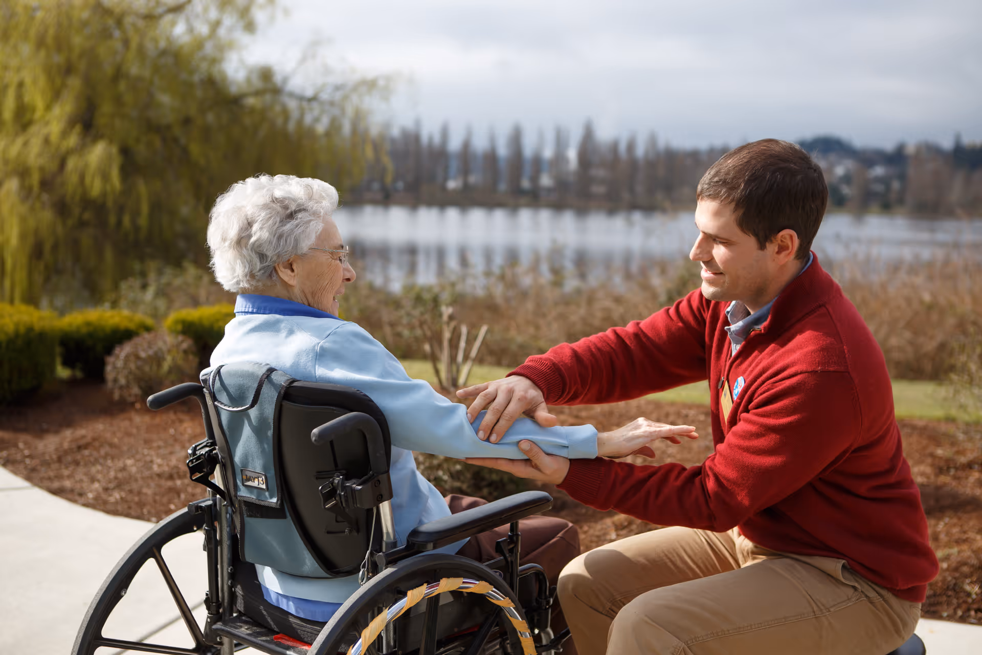 An elderly woman in a wheelchair is outdoors by a lake, accompanied by a man in a red sweater who is holding and stretching her arm, both smiling and engaging in a friendly interaction.