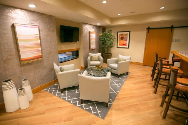 A cozy common area in a nursing home featuring four white armchairs arranged around a glass coffee table on a gray patterned rug. The room has wooden flooring, modern wall art, a flat-screen TV mounted on the wall, a potted plant, and a bar counter with stools on the right side.