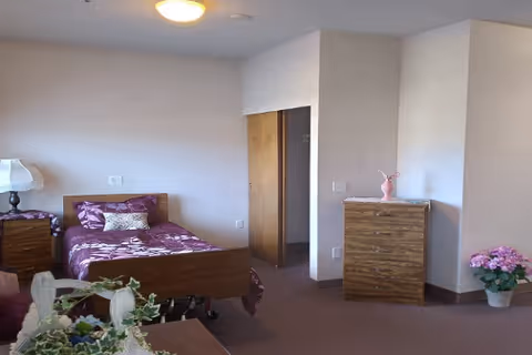 A simple bedroom in an assisted living facility with a single bed covered in purple floral bedding, a wooden nightstand with a lamp, a wooden dresser with a pink vase on top, and a potted plant with pink flowers on the floor. The walls are plain and light-colored, and there is an open doorway leading to another room.