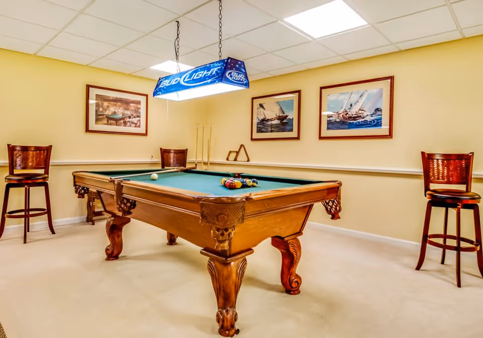 A well-lit room with a wooden pool table set up for a game, featuring a green felt surface and a rack of billiard balls. Above the table hangs a Bud Light branded light fixture. The room has beige walls adorned with framed pictures of sailboats and a pool hall scene. Two wooden bar stools are positioned against the walls on either side of the pool table.