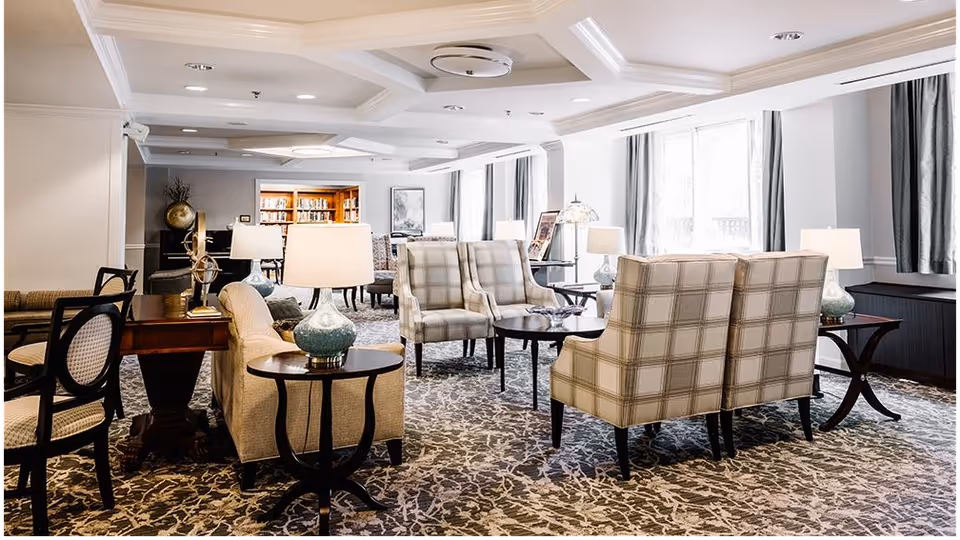 A bright and spacious living room area in a retirement community featuring multiple upholstered armchairs and sofas arranged around small tables with lamps. Large windows with curtains allow natural light to fill the room, and a bookshelf is visible in the background.
