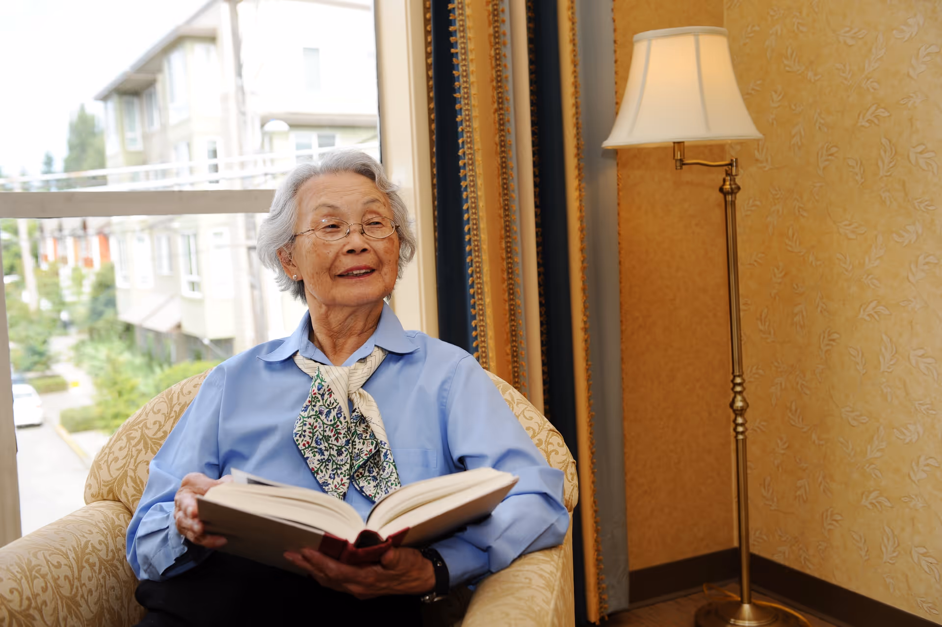 An elderly woman with gray hair and glasses sits in a patterned armchair near a window, holding an open book and looking to the side with a gentle smile. She is wearing a blue shirt and a floral scarf. Behind her is a tall floor lamp and a wall with a subtle floral pattern.