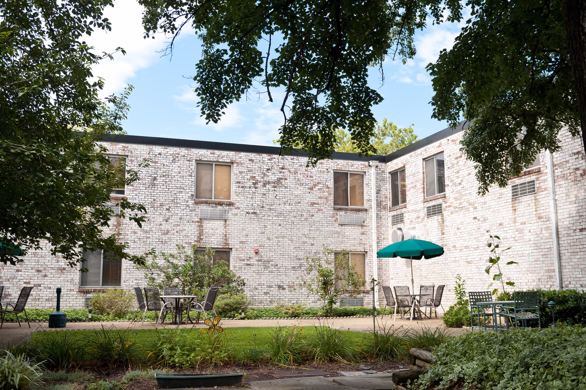 Outdoor courtyard area at Autumn Lake Healthcare at Pikesville featuring a brick building with multiple windows, several patio tables and chairs, one with a green umbrella, surrounded by greenery and trees.