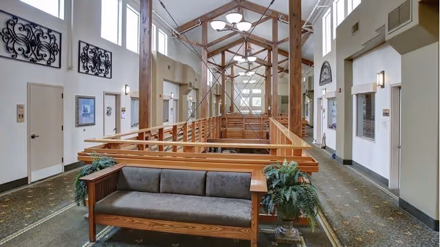 Interior view of a senior living facility hallway with high ceilings, wooden beams, and large windows. There is a wooden bench with gray cushions and potted plants on either side. The hallway has doors and framed artwork on the walls, with carpeted flooring and modern light fixtures hanging from the ceiling.