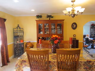 A dining room with a wooden dining table covered with a floral tablecloth, surrounded by wooden chairs. On the table is a vase with red and orange flowers and two red candles. Behind the table is a wooden china cabinet with glass doors displaying dishes and glassware. To the left is a metal shelving unit with decorative items and a globe. The walls are painted yellow with white trim, and a chandelier with multiple lights hangs above the table. In the background, there is a glimpse of another room with a person sitting in a wheelchair.