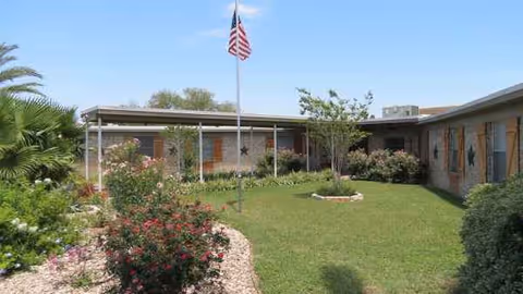 A single-story senior living facility exterior with a manicured lawn and flagpole in front.