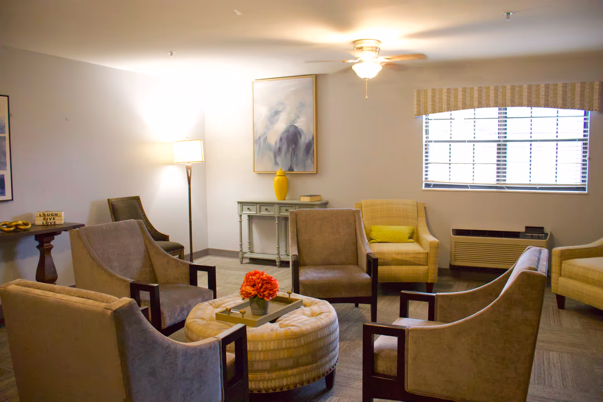 A cozy living room area with six armchairs arranged around a round ottoman with a tray and a flower arrangement on top. The room has a window with blinds and a valance, a ceiling fan with light, a floor lamp, a small console table with a yellow vase and a book, and framed artwork on the walls.