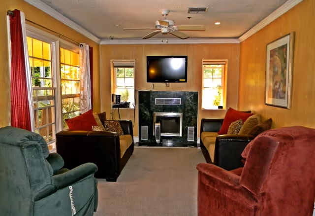 Cozy living room with upholstered chairs and sofas arranged around a fireplace and wall-mounted TV, with windows and a ceiling fan.