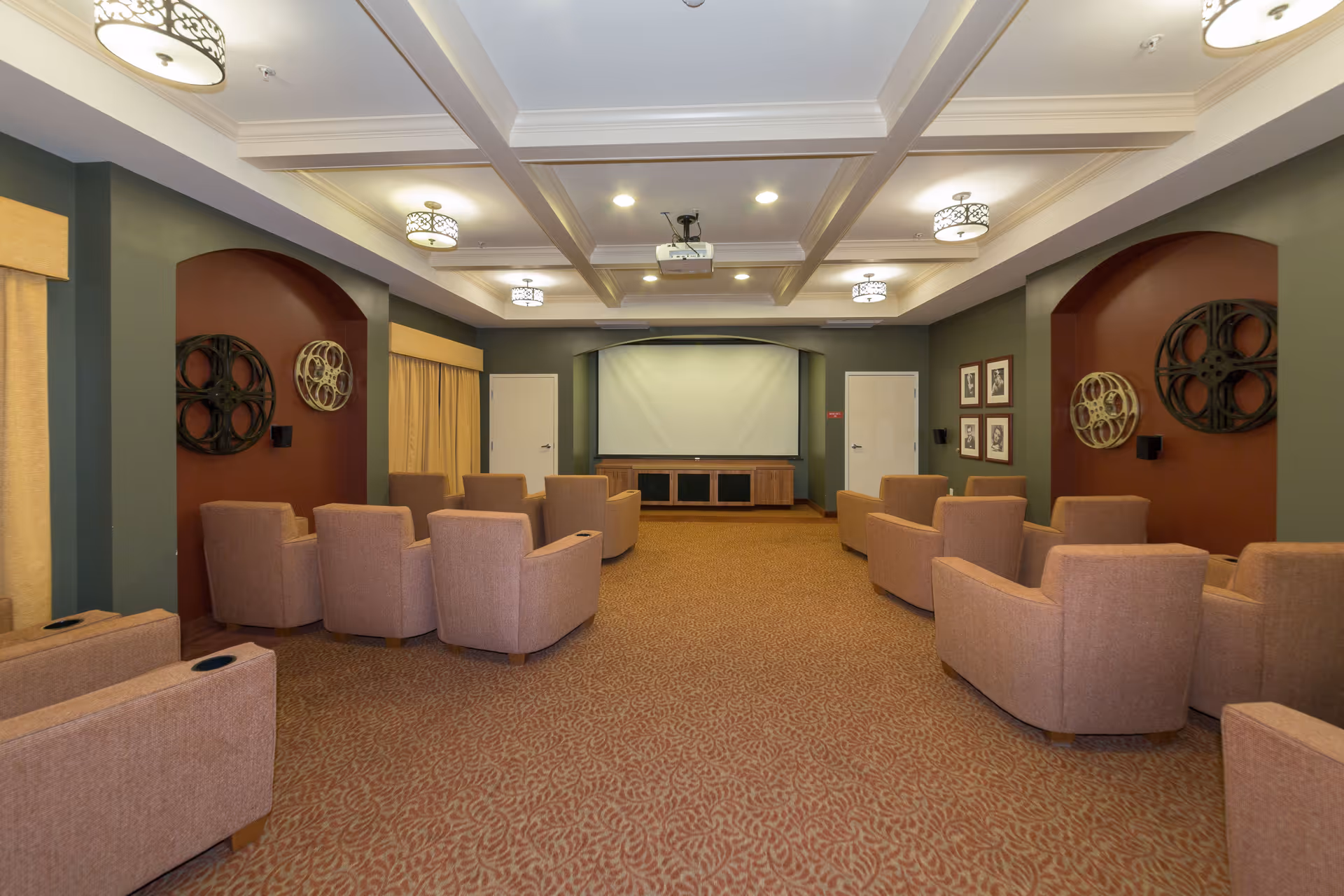 A cozy media room with multiple beige armchairs arranged in rows facing a large projector screen. The room has a patterned carpet, green and maroon walls with decorative film reel wall art, and ceiling lights with decorative covers.