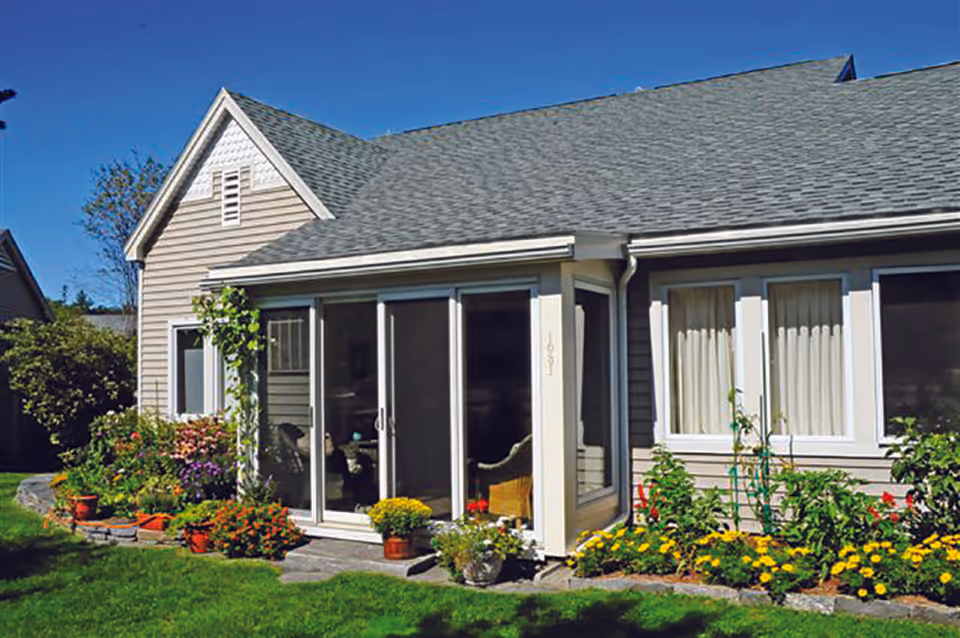 Exterior view of a single-story residential building with gray siding and a shingled roof. The building features large sliding glass doors and windows with white trim. There is a well-maintained garden with various colorful flowers and green plants along the foundation, and a green lawn in the foreground under a clear blue sky.