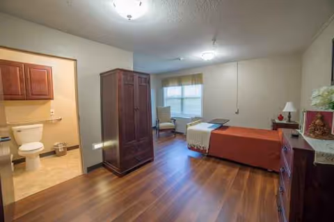 A simple bedroom in a senior living facility with a bed, dresser, and adjoining bathroom.